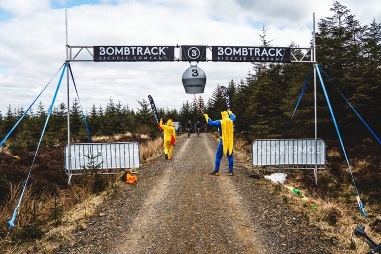 Banana Man and Chicken Girl cheer riders on under a Bombtrack banner on a forest track at Dirty Reiver 2019.