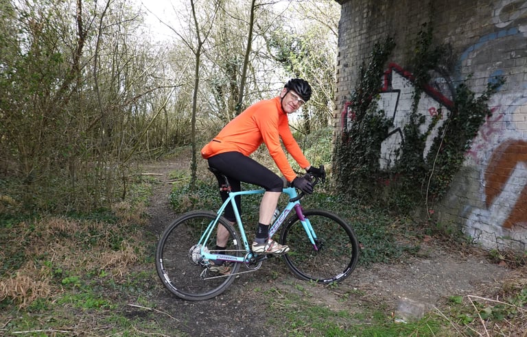 Rider in an orange jacket on the Orbea Terra beside a graffitied brick bridge on a rough path.
