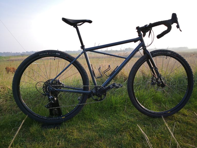 Side view of the grey BiViBIKES Bunker gravel bike in long grass, with flat open fields and a cow behind.