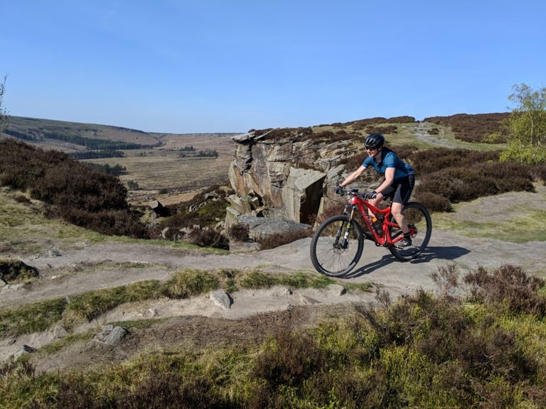 Rider in Alpkit Floe women's MTB shorts on a rocky moorland track past a gritstone outcrop above a wide valley.