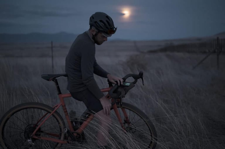 Rider at dusk under a low moon, stopped in long grass with a Wahoo ELEMNT ROAM on the handlebars of a gravel bike.