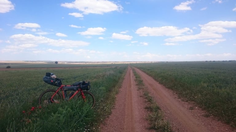 Red bikepacking bike with bar and saddle bags leans in a green field beside a long red-dirt track.