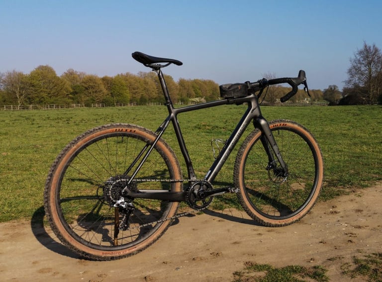 Bombtrack Hook EXT-C in satin black with tan-wall WTB Sendero tyres, parked on a dirt track in front of an open field.