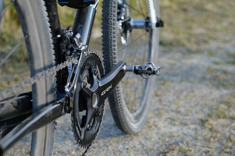 Close-up of a Shimano GRX crank and front derailleur, with a gravel tyre and pedal behind it, shot at a low angle.