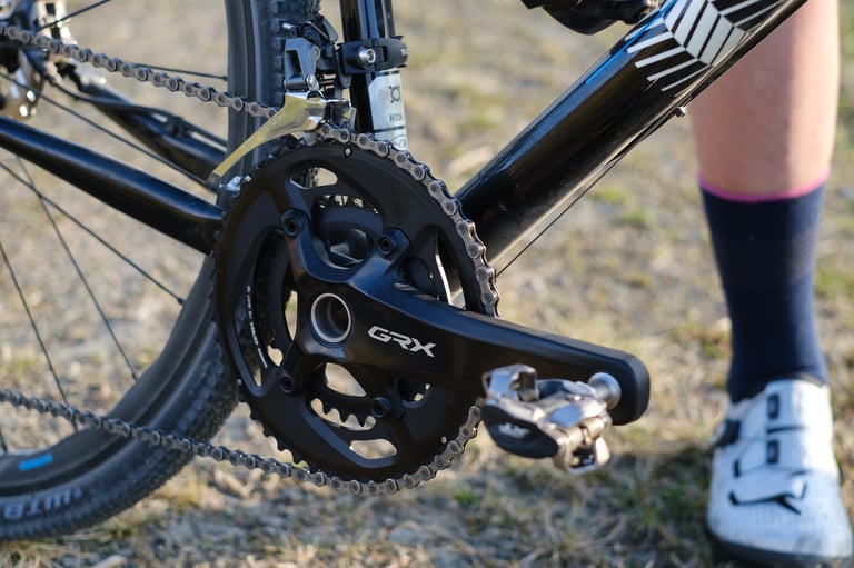 Shimano GRX double crankset with front derailleur and clipless pedal, a rider's shoe beside it on dry grass.