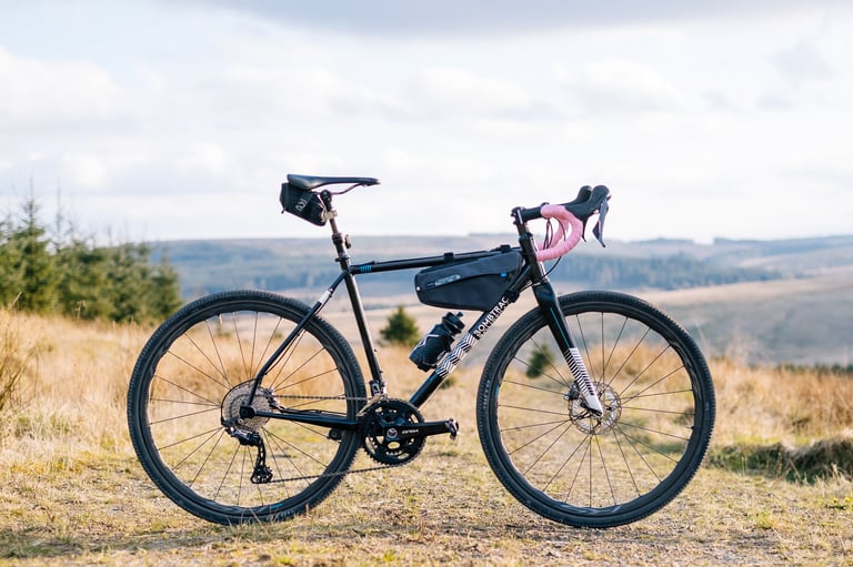 Black gravel bike with pink bar tape and Shimano GRX 2x drivetrain, parked on a grassy hillside above forest and moorland.