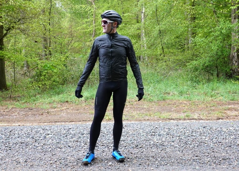 Rider standing on a gravel track in the black 7Mesh Oro jacket, tights and blue shoes, with woodland behind.