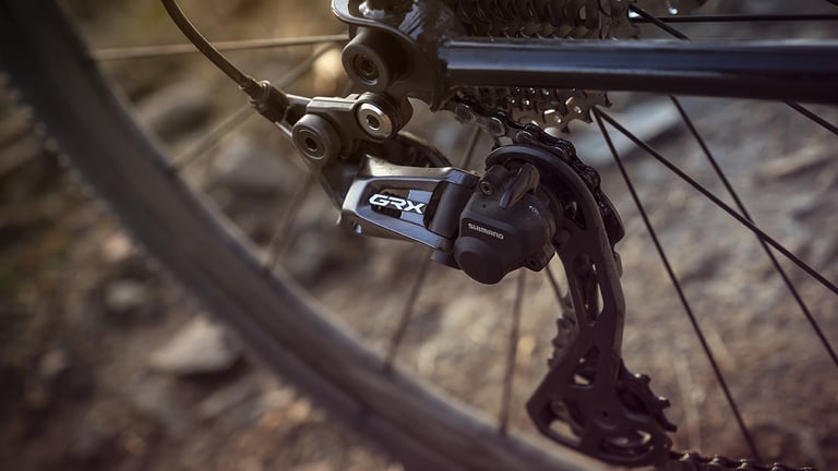 Shimano GRX rear derailleur with its long cage hanging below the cassette, set against a spoked wheel on rocky ground.