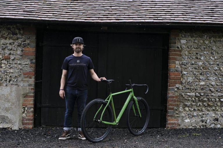 Dom Mason stands beside the green Bokeh 2 in front of the dark barn doors at the Sussex workshop.