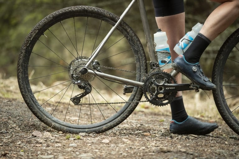 Shimano GRX gravel wheel and 2x drivetrain on a titanium bike on a forest track, a rider's foot resting on the pedal.