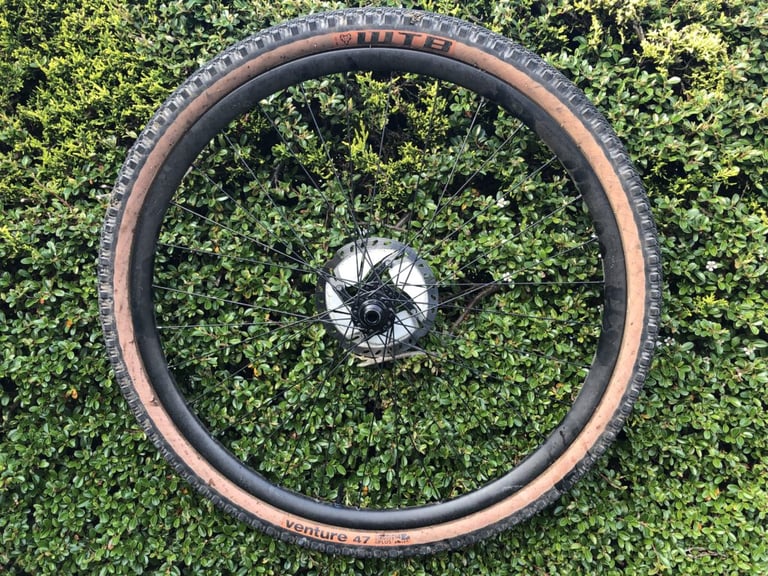 Whole front wheel with a tanwall WTB Venture 47 tyre, WTB logo uppermost, held against a hedge after testing.