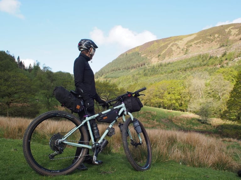 Rider beside a bikepacking bike on HUNT wheels with frame and bar bags, looking over a green valley.