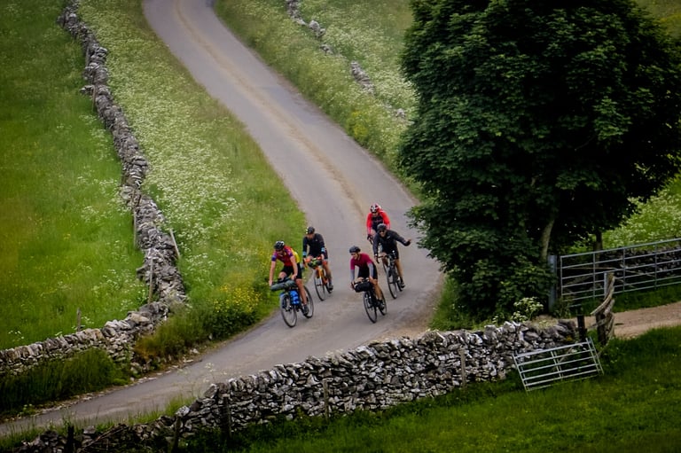 Five riders on gravel bikes with bikepacking bags roll down a quiet lane between dry stone walls on The Distance.