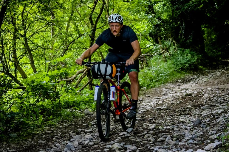 Rider with a handlebar bag on an orange hardtail mountain bike climbs a rocky trail through woodland at The Distance.