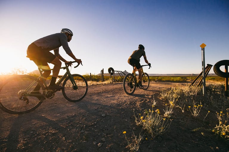 Two riders on Santa Cruz Stigmata gravel bikes roll along a dirt road through desert scrub in low sun.