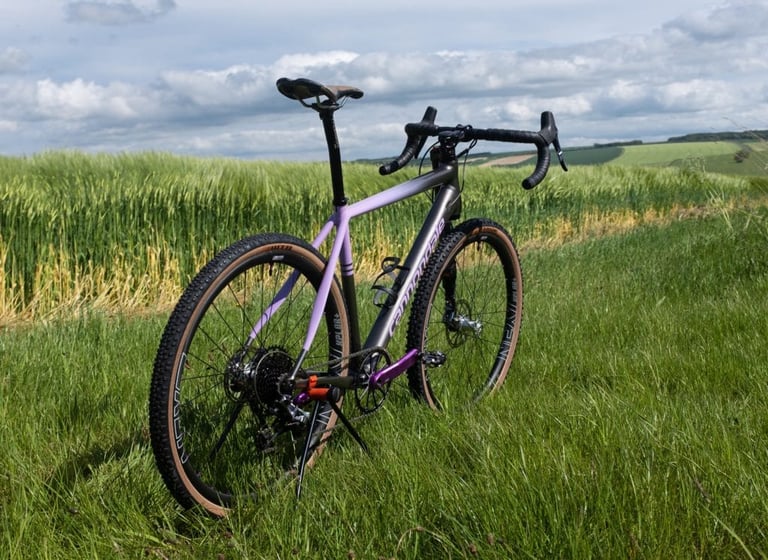 Rear three-quarter view of the Grinduro-painted Cannondale Slate in lilac and grey beside a green crop field.