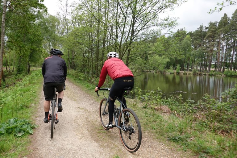 Two riders pass a still lake on a sandy forest track during the Gravel’duro preview ride at Swinley.