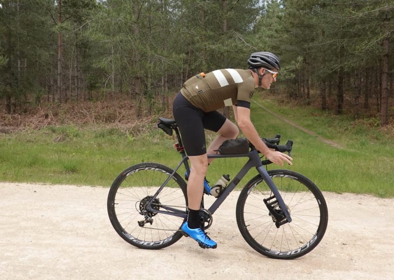 Rider in the Blackmore Cadet jersey pedals a gravel bike along a pale forest road lined with pines.