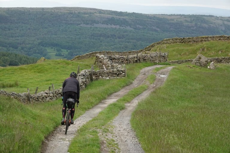 A rider on a loaded gravel bike descends a stony double track between drystone walls in the Lake District.