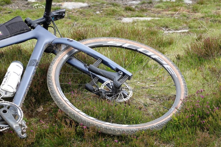 Front wheel of a muddy grey gravel bike with a Scope rim and tan-wall tyre lying in heather.