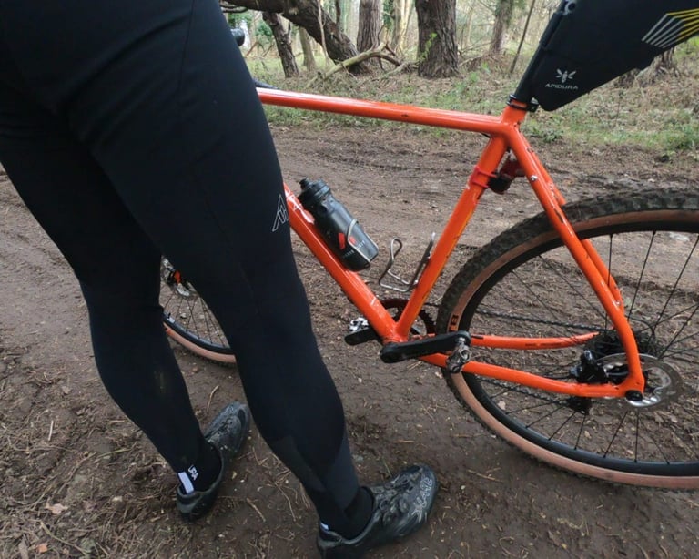 Rider in black tights beside an orange gravel bike with a tan-wall tubeless tyre on a muddy woodland track.
