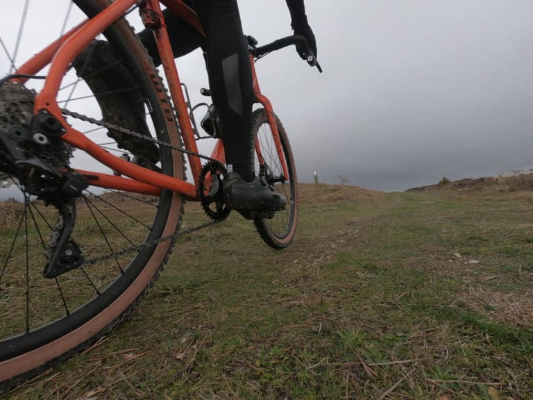 Low view of an orange gravel bike with a muddy tan-wall tyre on a grassy moorland track under grey cloud.