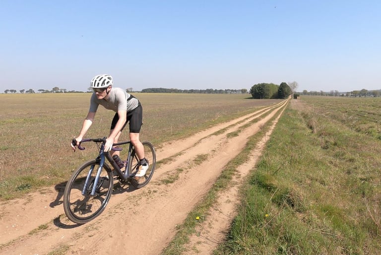 Rider in the Ashlu Merino Jersey heads towards the camera along a dry, sandy farm track under a blue sky.
