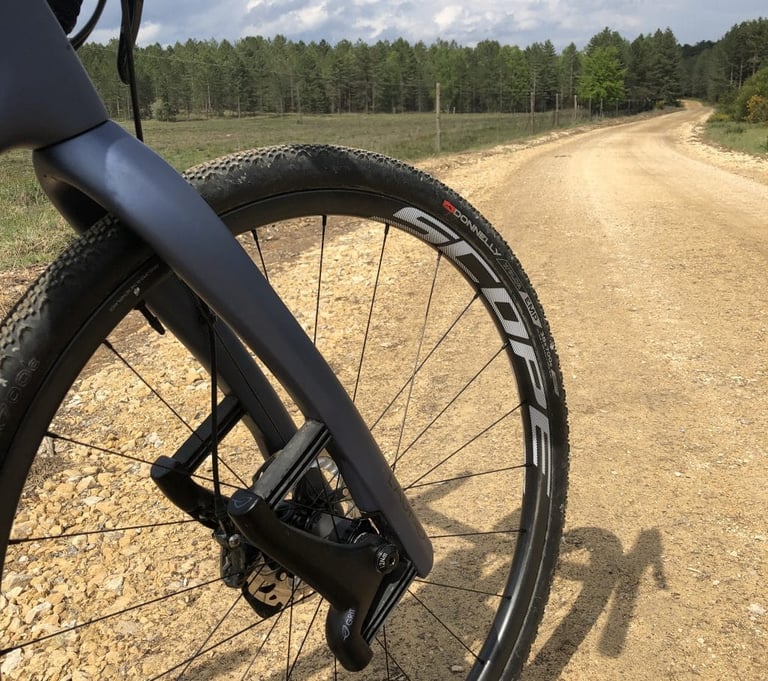 Donnelly EMP gravel tyre on a Scope wheel and carbon fork, on a wide stony fire road lined with pine woods.