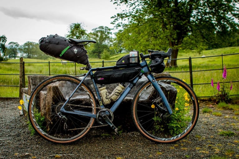 Blue Lauf True Grit with Miss Grape saddle pack, frame bag and top tube bag, leaning on a stone block by a fence.