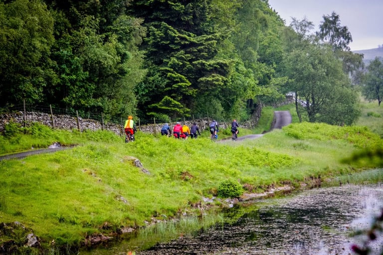 A group of riders with bikepacking bags rolls along a narrow Lake District lane beside a pond on The Distance.