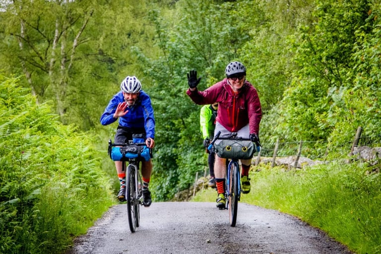 Two riders with handlebar bags wave to the camera on a wet, tree-lined Lake District lane during The Distance.