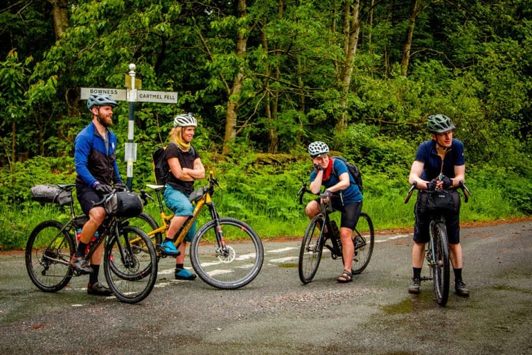 Four riders with loaded bikes pause at a signpost for Bowness and Cartmel Fell, checking the way on The Distance.
