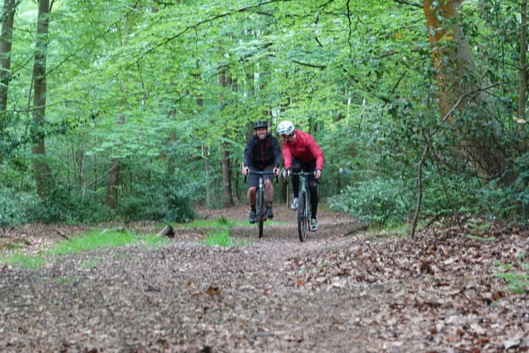 Two smiling riders approach on gravel bikes along a leaf-covered woodland trail in Swinley Forest.