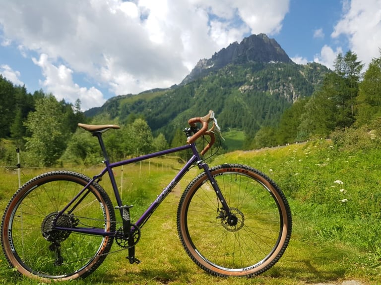 Velo Orange Piolet in purple standing on a grassy Alpine track with a rocky peak rising behind.