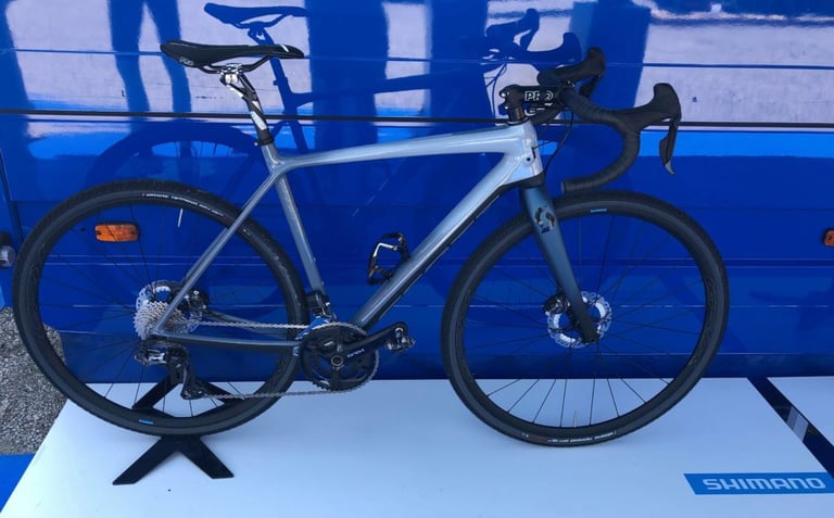 Side view of the grey gravel bike with a full Shimano GRX Di2 groupset on a display stand beside a Shimano sign.