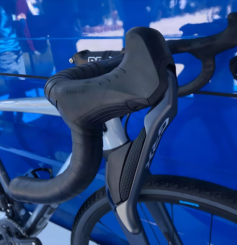 Shimano GRX ST-RX815 Di2 shifter with raised hood on gravel drop bars, against a blue Shimano display backdrop.