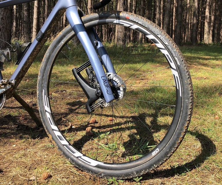 Scope O2 front wheel with a Donnelly tyre fitted to a Lauf gravel fork, parked on grass among pine trees.