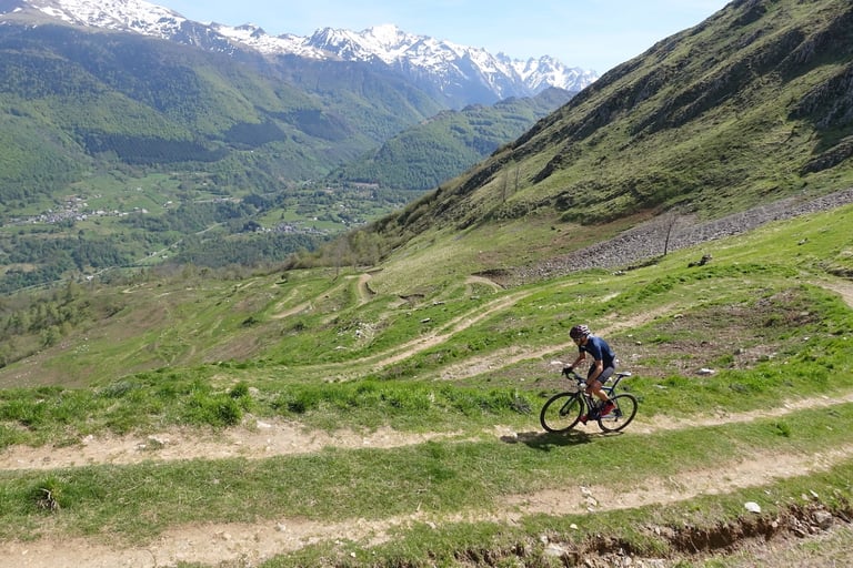 Gravel cyclist on a zigzagging hillside track in the High Pyrenees, with a valley village and snowy peaks beyond.