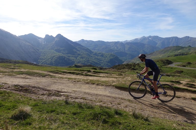 Rider on a blue gravel bike climbs a rocky dirt track above a green Pyrenean valley, with high peaks in the distance.