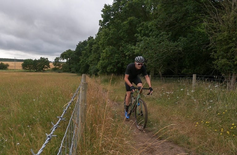 Gravel rider in PEdALED Odyssey kit follows a narrow dirt path beside a barbed-wire fence and long summer grass.