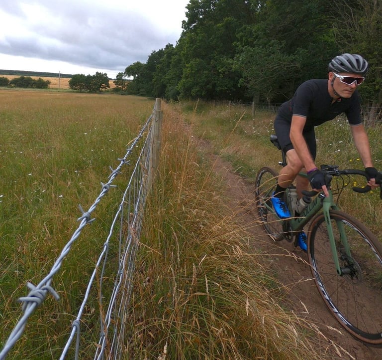 Rider on the Cipollini MCM All Road follows a dirt path beside a barbed wire fence through long grass.