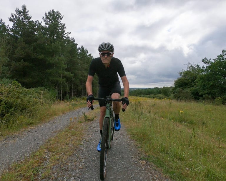 Rider in the PEdALED Odyssey jersey and bibshorts on a green gravel bike, riding a gravel track beside pine woodland.