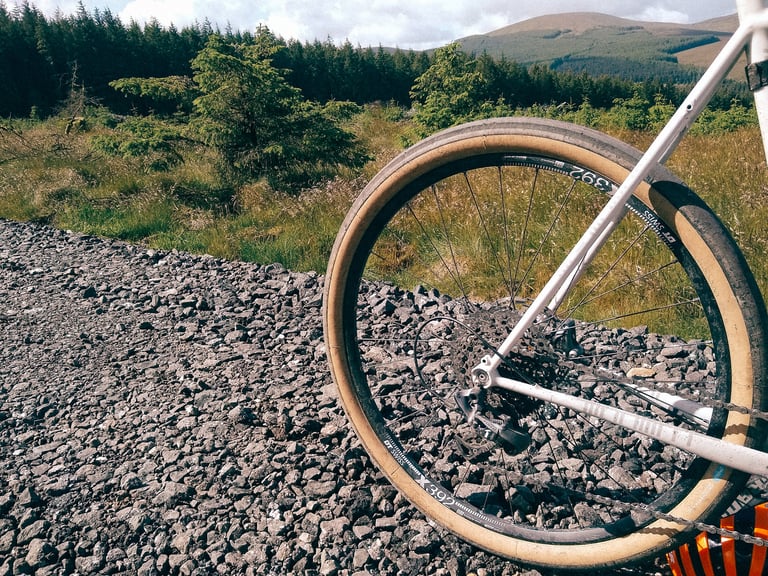 Rear wheel of a white gravel bike on the Switchback Hill tyre beside a stony forest track.