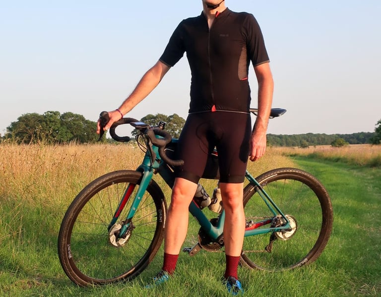 PEdALED Odyssey jersey front view, with mesh underarm panel, worn beside a teal gravel bike in a summer field.