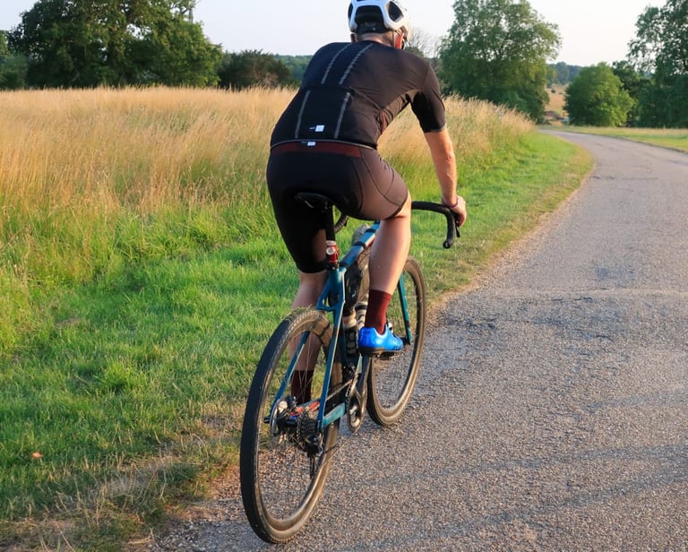 Rider in PEdALED Odyssey bibshorts and jersey rides away on a narrow tarmac lane at sunset, large rear pocket visible.