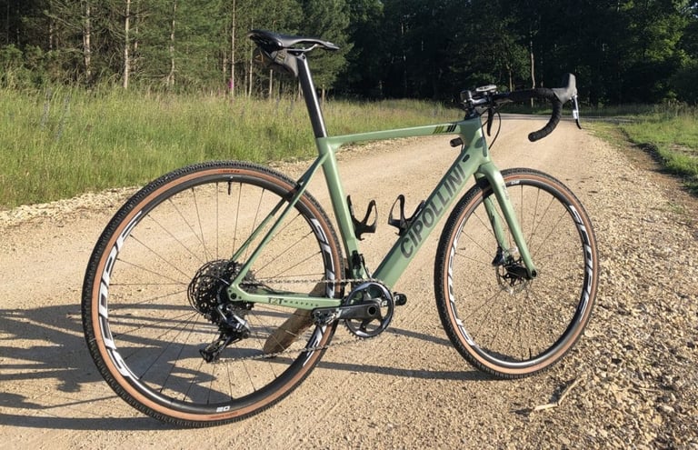 Scope O2 wheels on a green Cipollini gravel bike on a pale gravel track beside woodland.