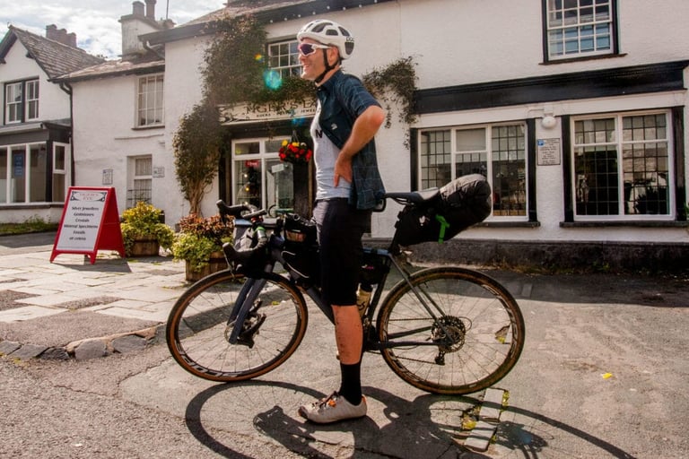Rider in a Morvelo Overland shirt beside a loaded gravel bike in a Lake District village.