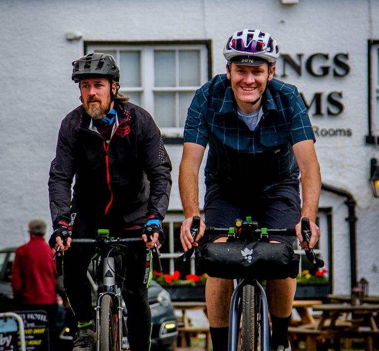 Two bikepackers ride past a pub, one wearing the Morvelo Overland check shirt, on a Lake District trip.
