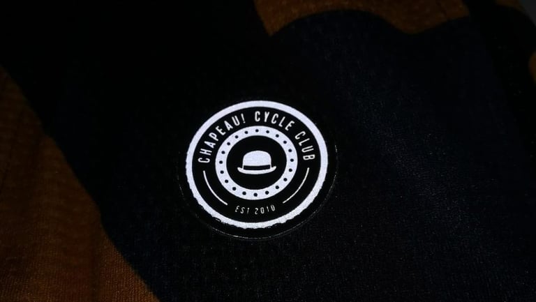 Small round reflective Chapeau Cycle Club badge on the black rear pocket of the Club Jersey Pro.