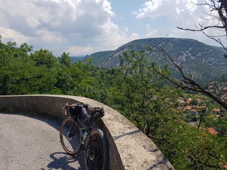 Loaded Velo Orange Piolet leaning on a stone wall at a hairpin, with wooded hills, a limestone ridge and red roofs beyond.